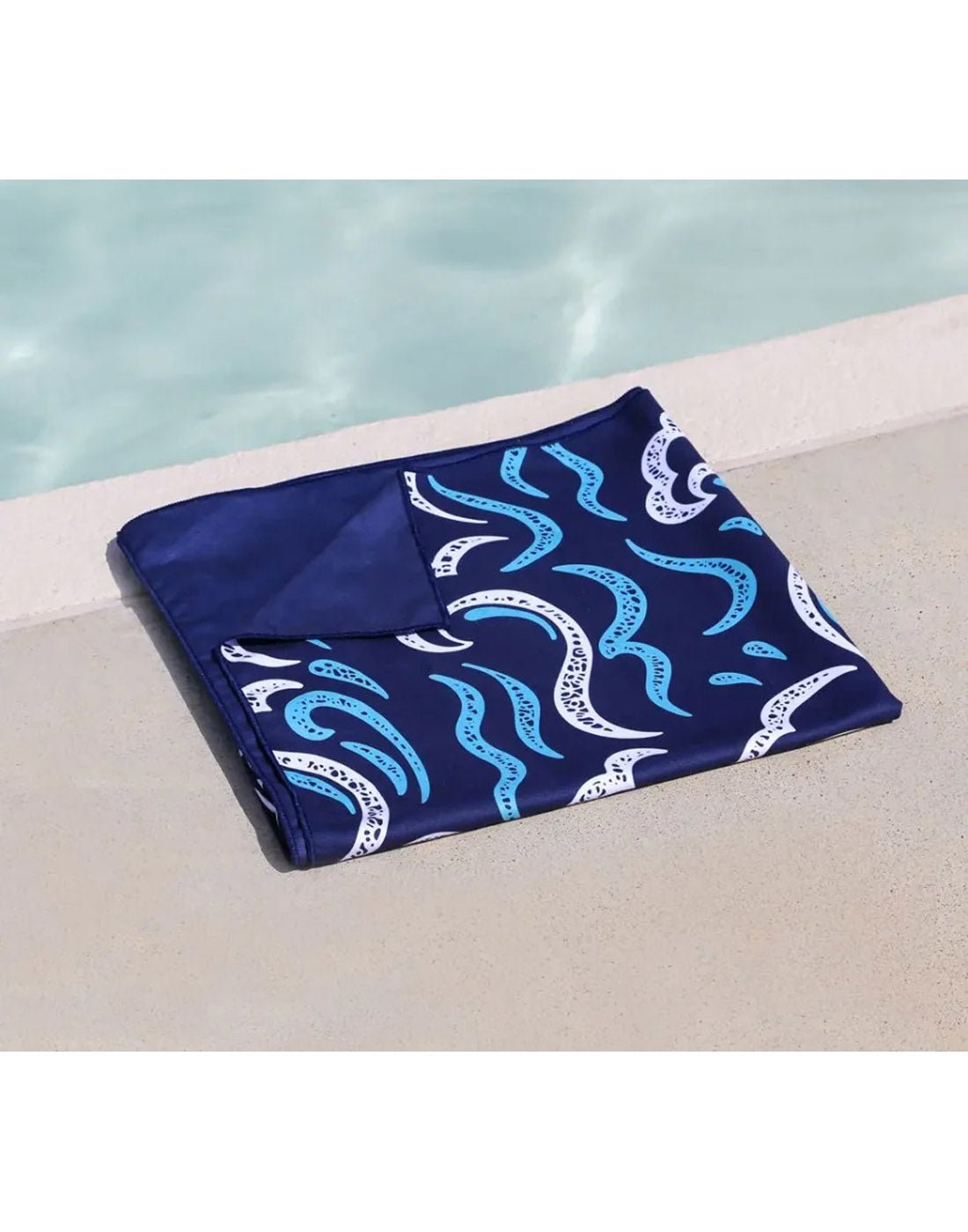 Lifestyle image of ocean K5 Microfibre Beach Towel, folded and positioned on the ground beside a pool.