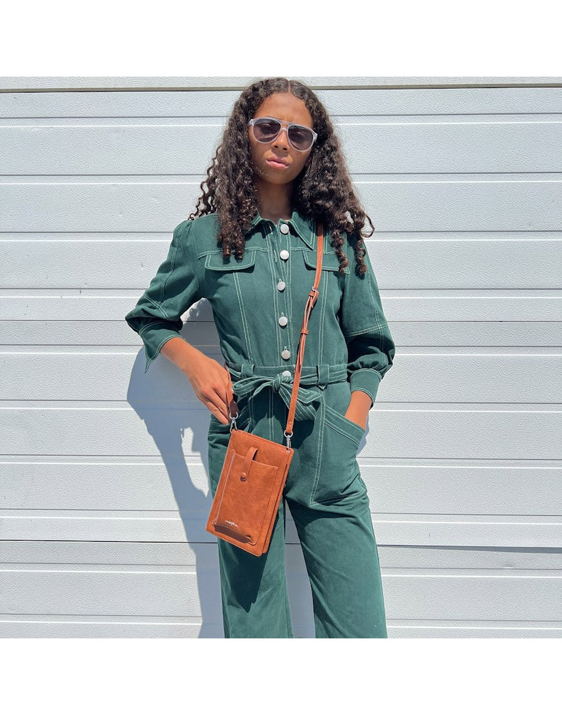 Woman wearing green jumpsuit and the Espe Pastel iSmart Pocket in tan as a crossbody while standing in front of a grey exterior wall