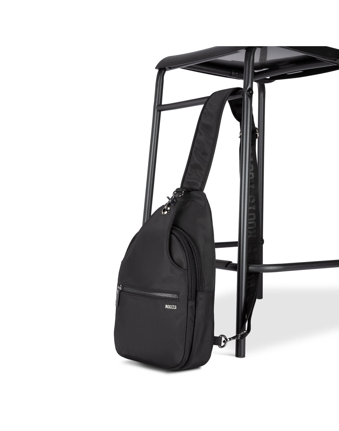 Side angle view of Roots Travel Sling Bag in black, positioned against a stool.