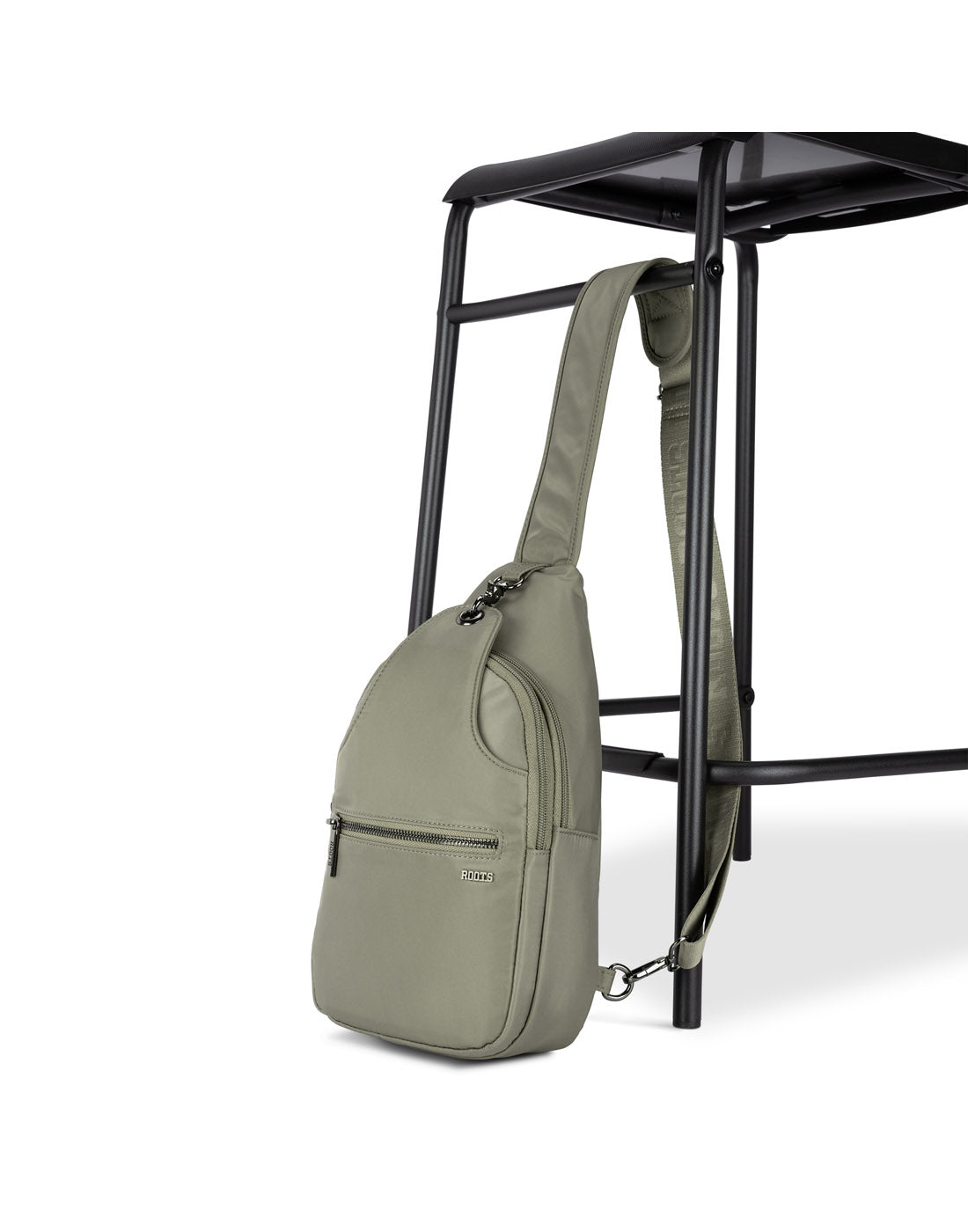 Side angle view of Roots Travel Sling Bag in sage, positioned against a stool.