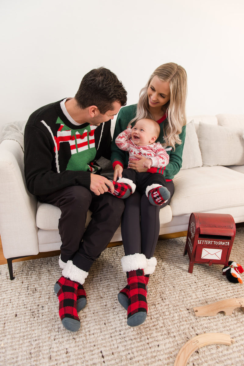 Man, woman and baby sitting on a couch wearing Christmas sweaters and Pudus Classic Slipper Socks in Lumberjack Red