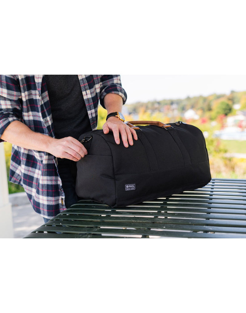 Man wearing open flannel shirt unzipping the PKG Bishop II Duffle Bag on a metal table