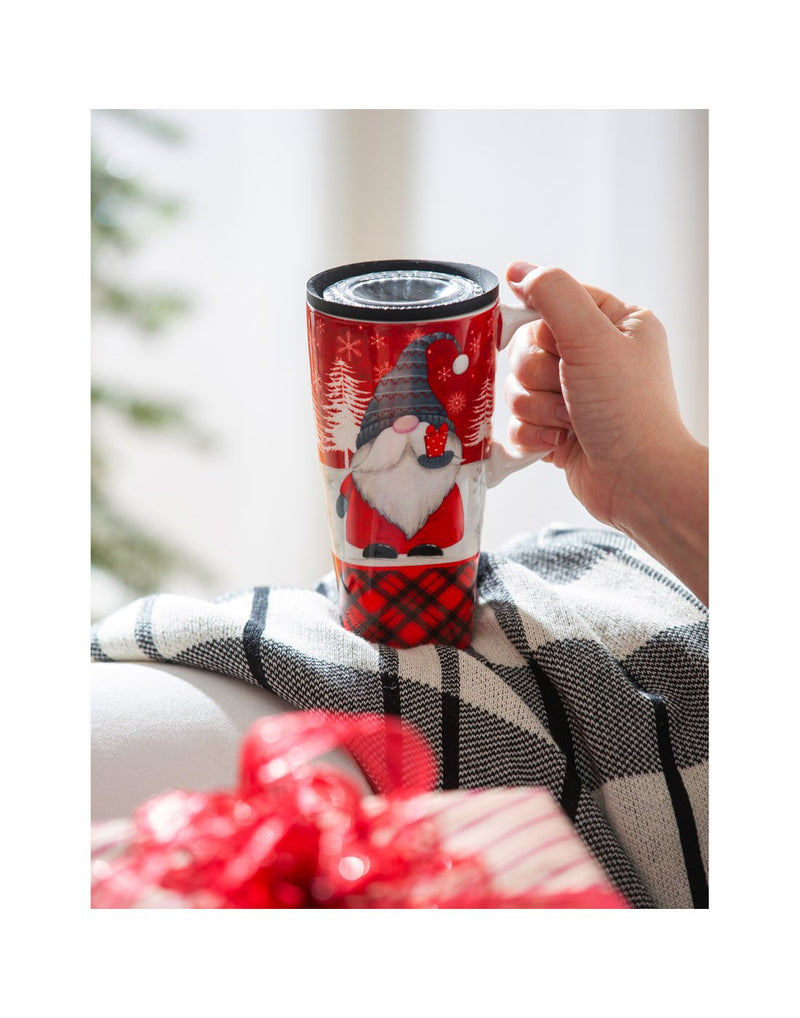 Hand holding the Ceramic FLOMO 360 Travel Cup - 17 oz Winter Gnome with white/black plaid blanket underneath and present in forefront