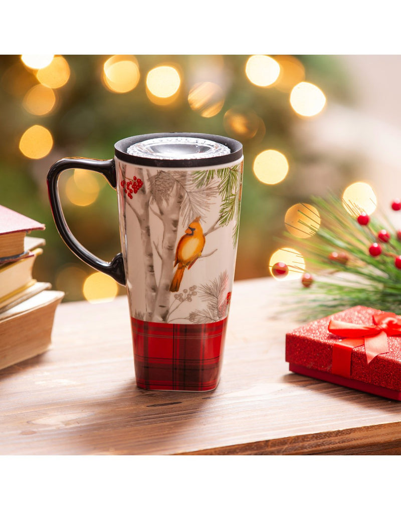 Ceramic FLOMO 360 Travel Cup - Christmas Cadence design with redish yellow cardinal on birch tree - cup is sitting on a wood table with books, small red present and berry branch beside and Christmas lights in background