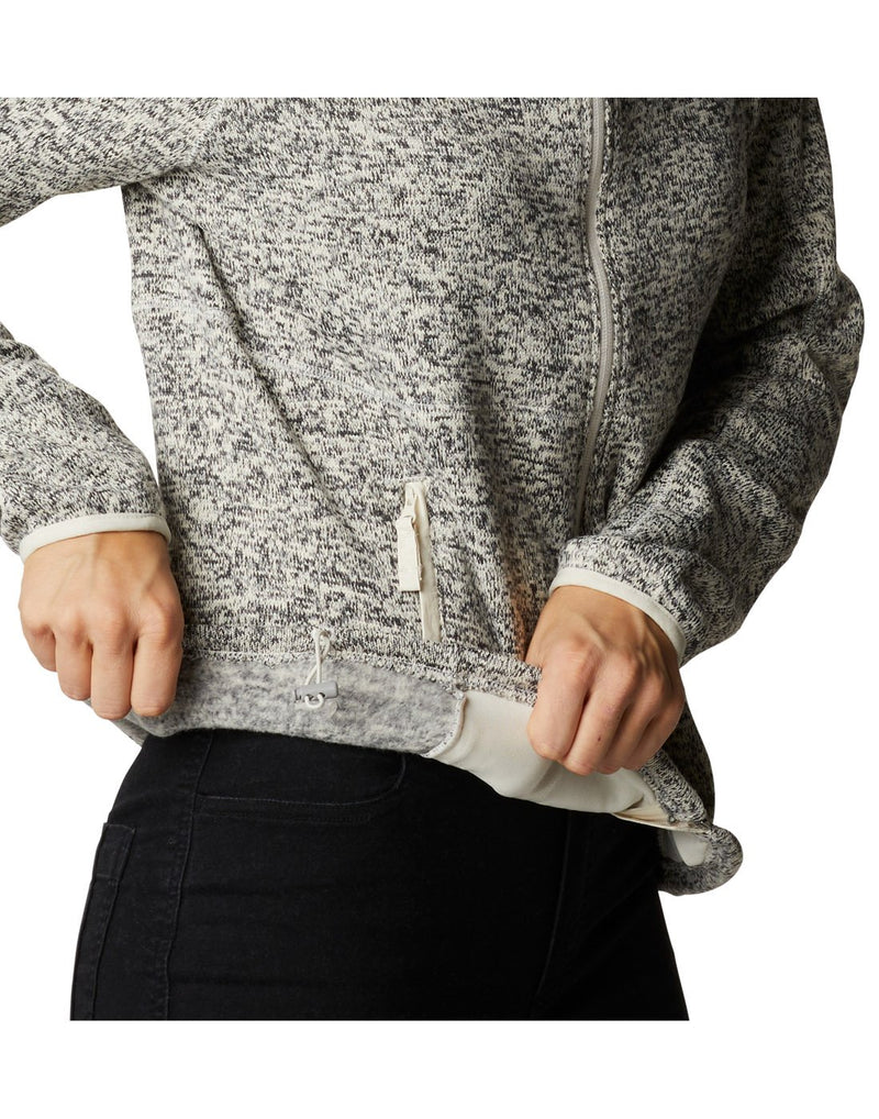Close up of woman wearing Columbia Women's Sweater Weather™ Full Zip Jacket in chalk heather, zipped up, side view holding up bottom hem to show adjustable drawstring