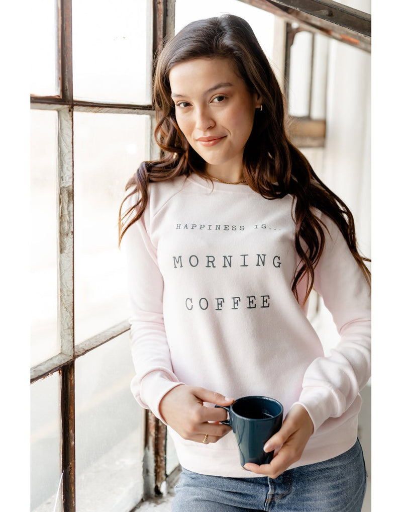 Woman standing by a window wearing blue jeans and Happiness Is... Women's Morning Coffee Crew Sweatshirt in ballet pink, holding a cup of coffee