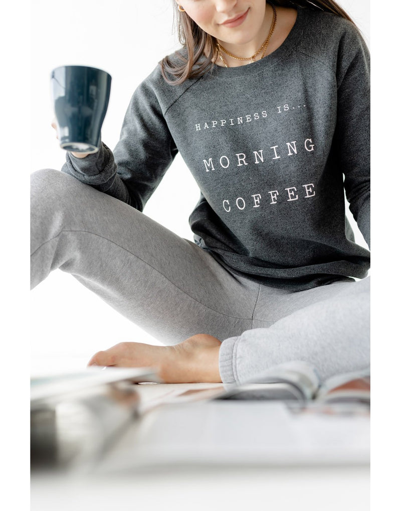 Woman wearing grey sweatpants and Happiness Is... Women's Morning Coffee Crew Sweatshirt in charcoal, holding a cup of coffee while sitting on the floor looking at a magazine