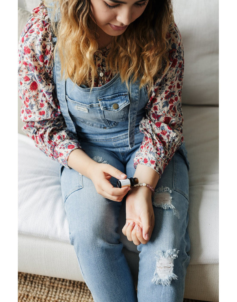 Woman wearing floral shirt and denim overalls, sitting on a white couch dropping essential oils on the beads of the Fern & Petal White-Howlite Bracelet