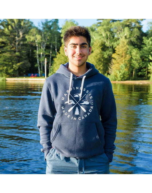 Product Image – Man wearing blue shorts and Happiness Is...Unisex Crest Hoodie in navy, with his hands in his pockets and a lake and trees behind him