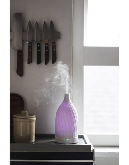 Fern & Petal White Ceramic Ultrasonic Diffuser with purple light and mist on, sitting on a kitchen counter with a window and knives in the background