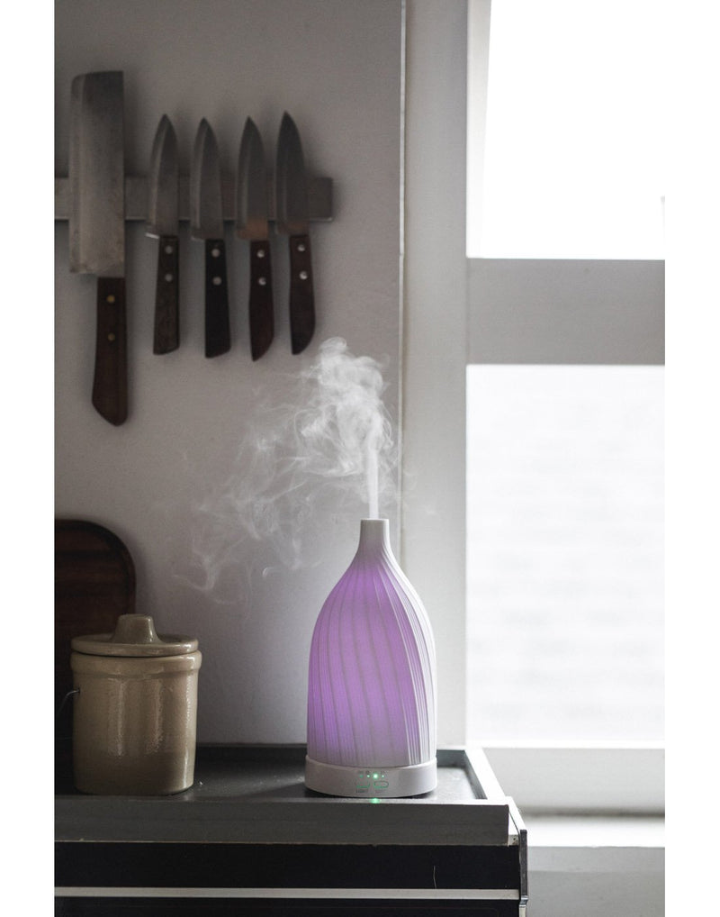 Fern & Petal White Ceramic Ultrasonic Diffuser with purple light and mist on, sitting on a kitchen counter with a window and knives in the background