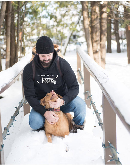 Product Image – Man wearing blue jeans, a black toque, a backpack, and Happiness Is...Hockey Unisex Hoodie in jet black crouched down on a bridge in a snow covered forest petting a brown dog