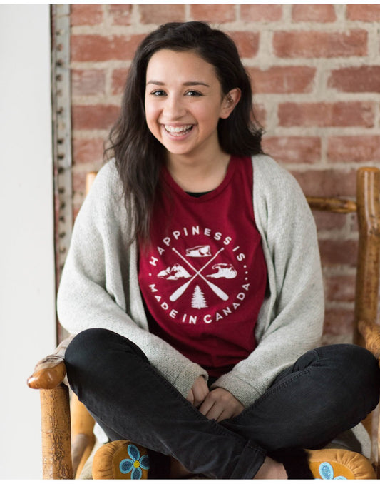 Product Image – Woman sitting cross-legged on a wooden chair wearing black jeans and a Happiness Is...Women's Crest T-Shirt in Canada Red with a light grey cardigan open on top