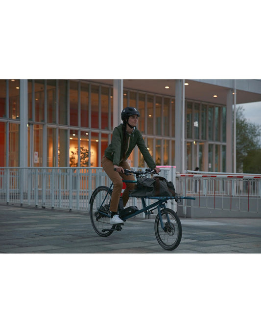 Product Image – Woman wearing the Briko E-One LED Reflective Bike Helmet riding a bike on a sidewalk infront of a building with long glass windows