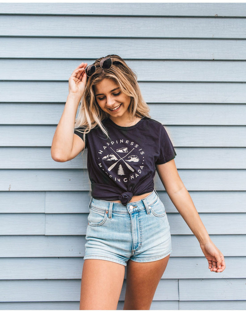Woman standing in front of a light blue siding wall wearing light blue jean shorts and a Happiness Is...Women's Crest T-Shirt in navy tied at the front, reaching for sunglasses on her head