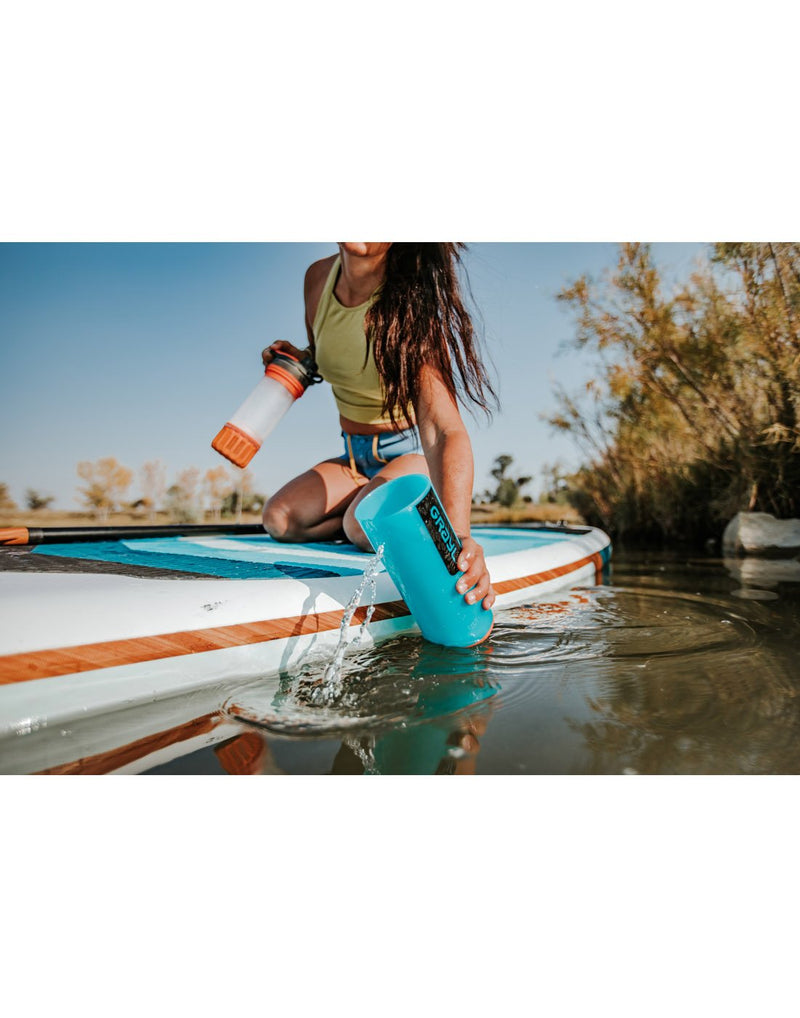 Woman scooping water into Grayl Geopress™ Water Purifier from side of a paddle board