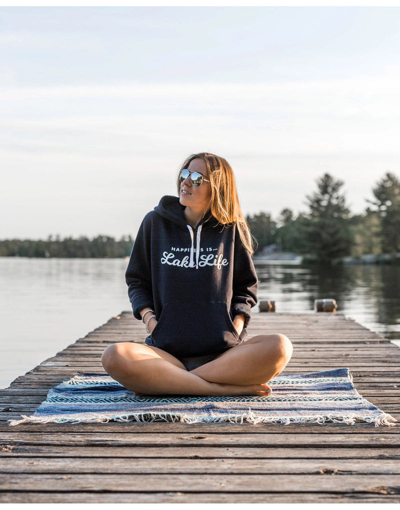 Woman wearing sunglasses and Happiness Is...Lake Life Unisex Hoodie in navy sitting cross-legged on a blanket on a dock with lake behind her