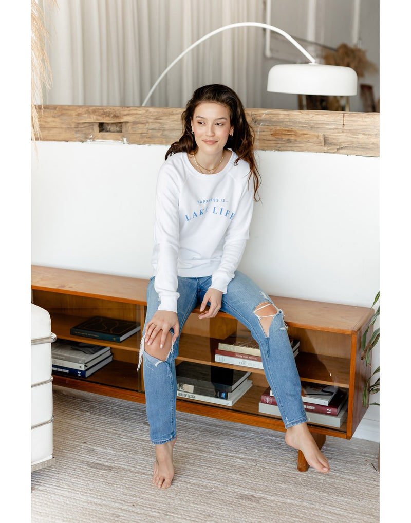 Barefoot woman wearing ripped blue jeans and white Happiness Is...Lake Life Women's Crew Sweatshirt sitting on a low bookshelf
