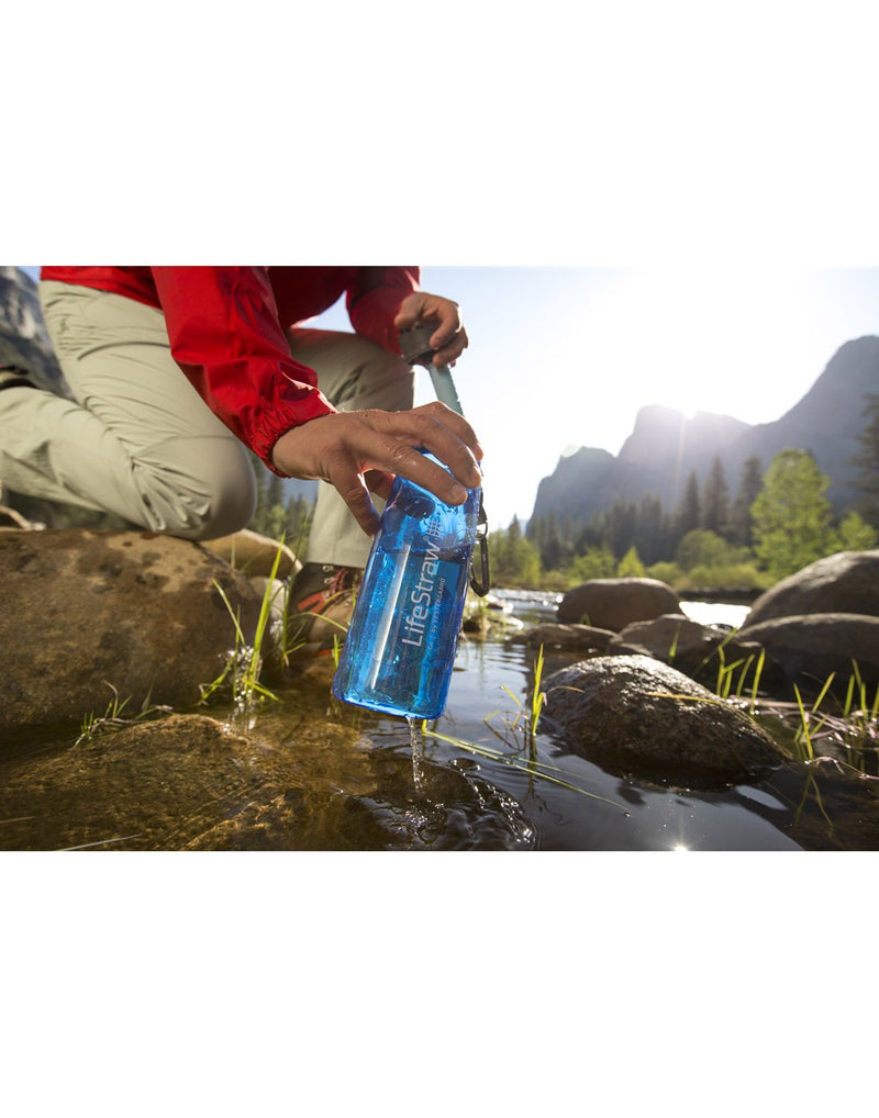 Person crouched down near rocks scooping up water in the blue LifeStraw Go Water Filter Bottle