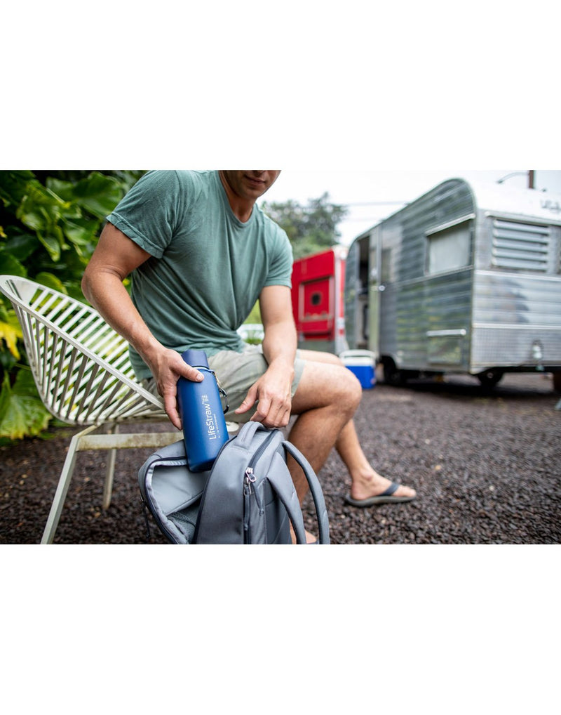 Man wearing shorts, t-shirt and sandals sitting on a white chair outdoors putting the blue LifeStraw Go Stainless Steel Water Filter Bottle into a grey backpack with a camper in the background