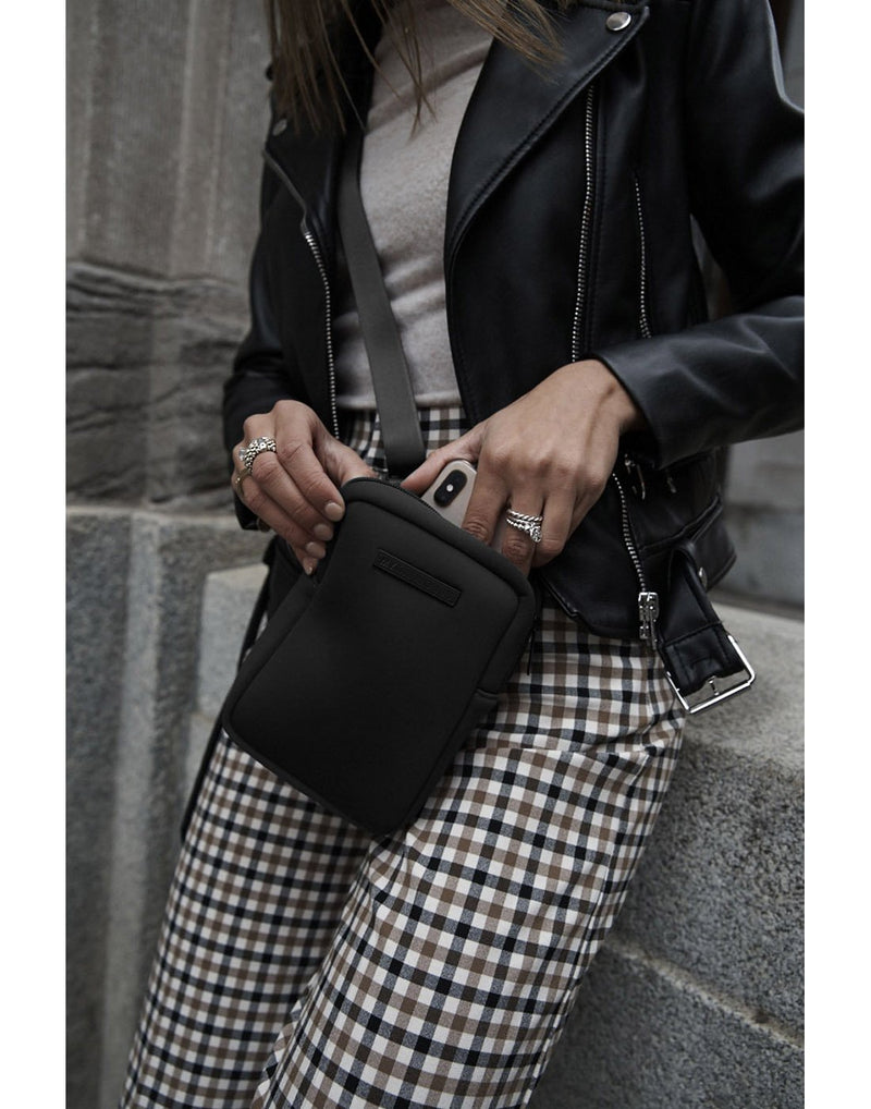 Woman wearing leather jacket and checkered pants taking a phone out of the MyTagAlongs mini crossbody that she has across her front