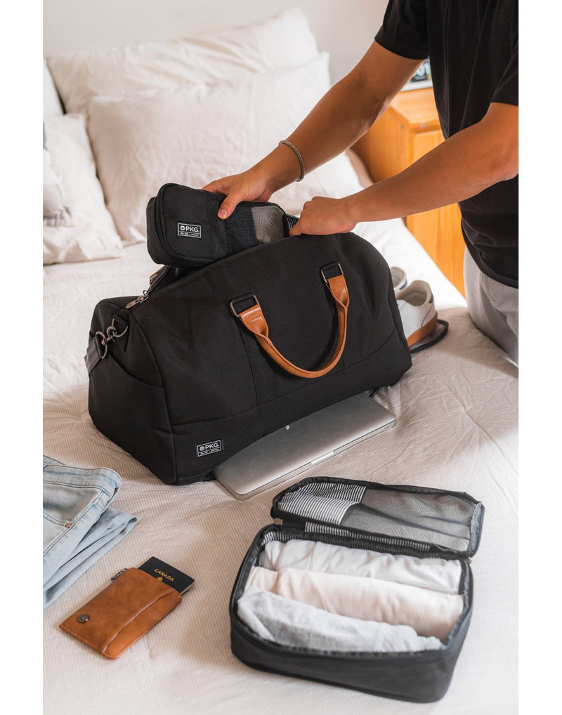 Man packing the PKG Bishop II Duffle Bag on a bed with Union packing cubes, clothes and passport around