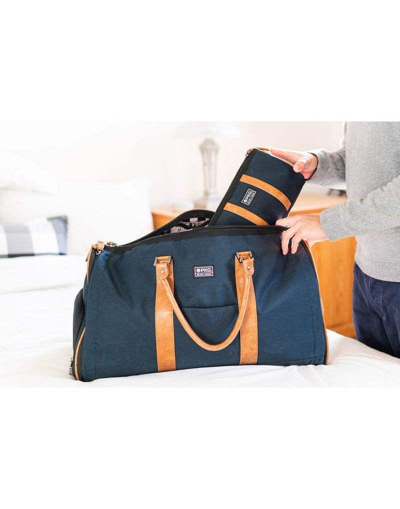 Person packing the navy PKG Charlotte Dopp Kit into a navy duffle bag on a bed