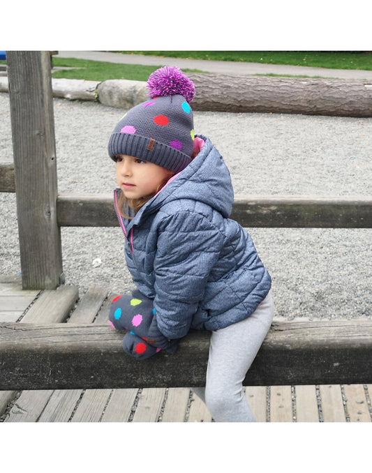 Product Image – Child in a grey winter coat sitting on a wooden fence wearing Pudus Kids Toque Winter Hat and mittens in Polka Dot Multi