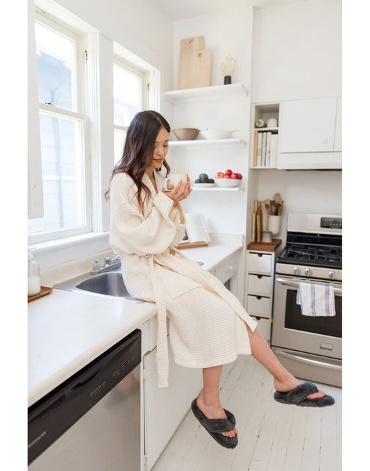 Product Image – Woman wearing white robe and grey Pudus Cottontail Flip Flop Slippers holding a cup of coffee while sitting on a kitchen counter