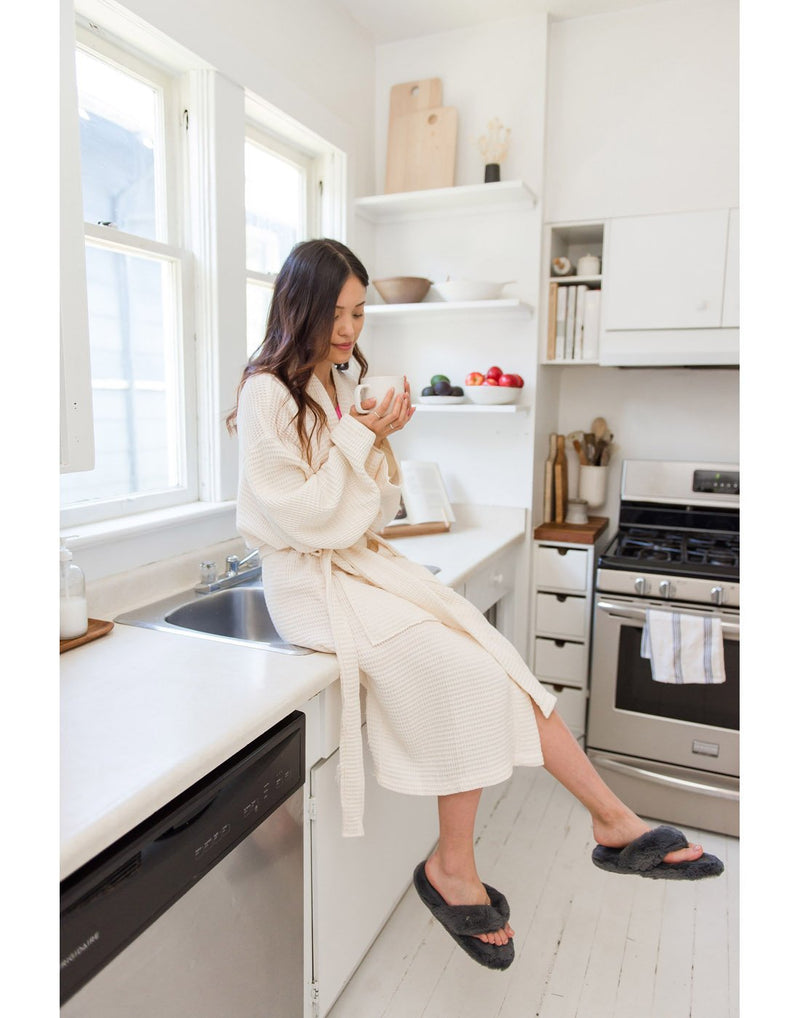 Woman wearing white robe and grey Pudus Cottontail Flip Flop Slippers holding a cup of coffee while sitting on a kitchen counter