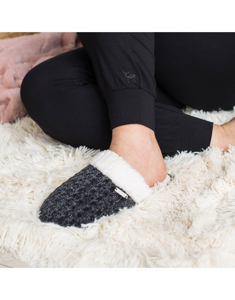 Close up of grey Pudus Creekside Chenille Knit Slide Slippers being worn by someone wearing black pants sitting on a white shag carpet