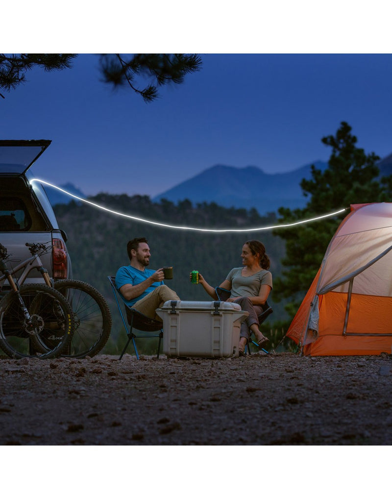 Nite Ize Radiant® Rechargeable Shineline™ set up between a parked car and a tent with two people sitting beneath having a drink, cooler and two bikes nearby