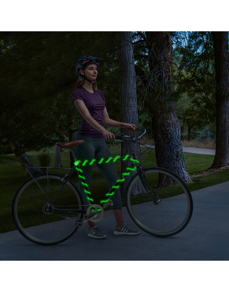 Person standing beside a bike on a path at dusk with the green Nite Ize Radiant® Rechargeable Shineline™ wrapped around the bike frame