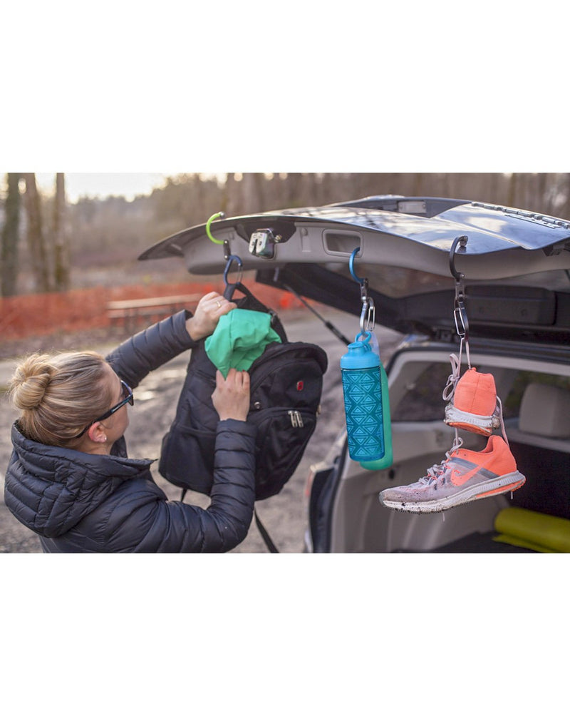 Woman wearing black winter jacket taking something out of a backpack that is attached with the small Heroclip to the open trunk of an SUV.  A pair of running shoes and a water bottle are also attached to the open trunk with separate small Heroclips