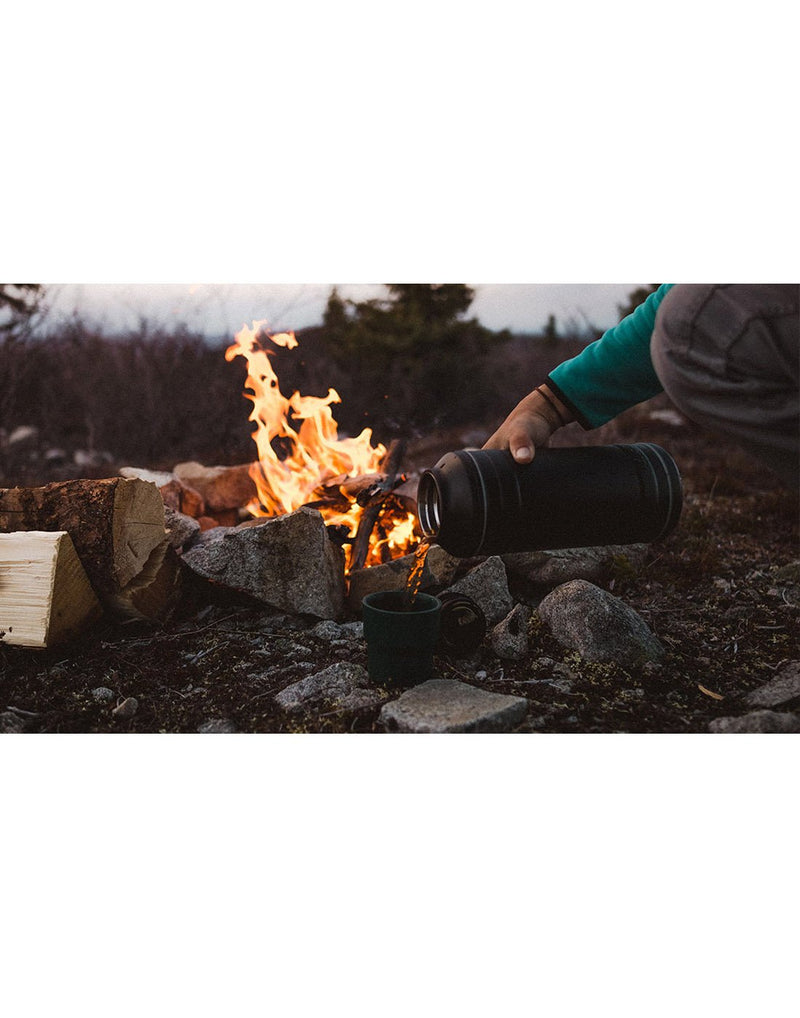 Someone pouring coffee from the black Pelican 64oz Bottle into a cup resting on some rocks on the ground infront of a campfire