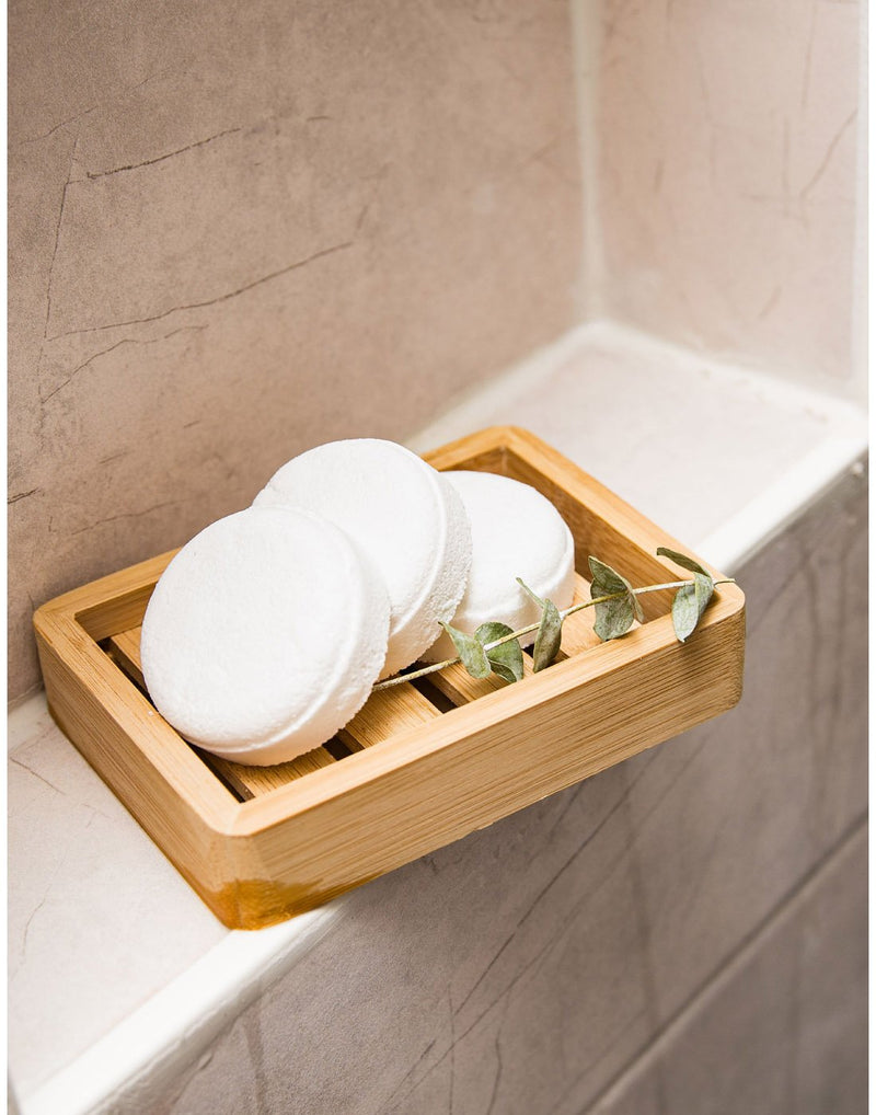 Three Bee by the Sea shower steamers with a sprig of eucalyptus on a wooden soap dish on a shower ledge